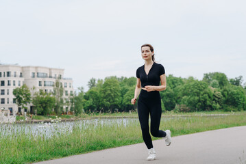 Fototapeta premium young slim beautiful caucasian woman running on path on river embankment in city park, black sportswear, green trees and grass, warm summer day