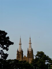 enchanting view of Jakarta Cathedral's twin spires and Gothic architecture peeking through lush green trees against a clear blue sky.