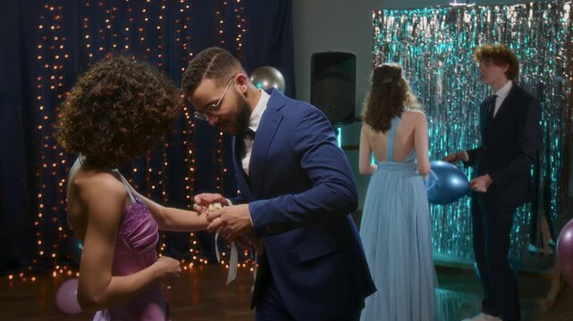 Young man in navy suit carfully attaching corsage to wrist of girl as another couple standing in the backgound at high school prom