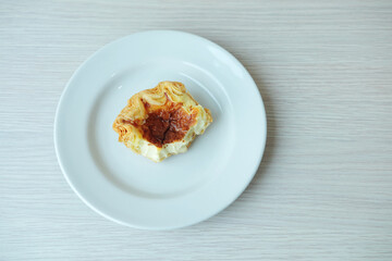 Half eaten custard pastry with golden flaky crust on a white plate over clean wooden table, minimal top view concept for snack, dessert, coffee break, or leftover food theme.