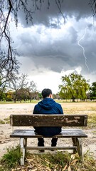 Person sits alone on bench in park under stormy sky