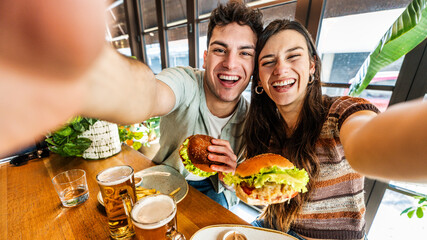 Fototapeta na wymiar Happy couple taking selfie with smart mobile phone at burger pub restaurant - Young people having lunch break at cafe bar venue - Life style concept with guy and girl hanging out on weekend day