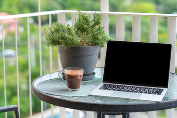Close-up of laptop and glass of hot chocolate on round balcony table with green plant. Concept of...