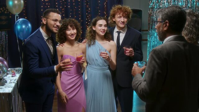 Group of young people in evening dresses and suits talking to their teachers while celebrating graduation at high school prom party