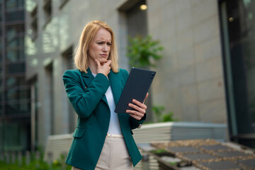 Thoughtful businesswoman in green blazer using tablet outdoors, professional woman analyzing data in modern urban environment  

