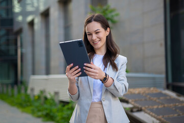 Fototapeta premium Smiling young businesswoman in blazer using tablet outdoors in modern urban environment, positive digital lifestyle concept 