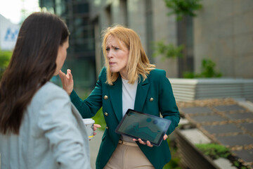 Two businesswomen arguing outdoors, frustrated professional holding tablet with charts during...
