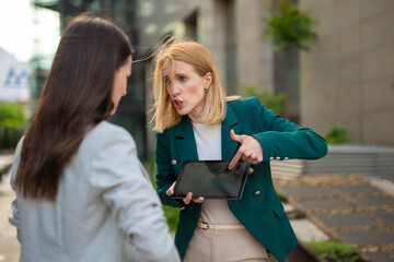 Frustrated businesswoman pointing at tablet while arguing with colleague outdoors in modern urban...