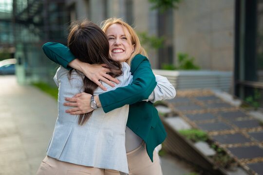 Two happy businesswomen hugging outdoors in modern urban environment, celebrating success and teamwork  
