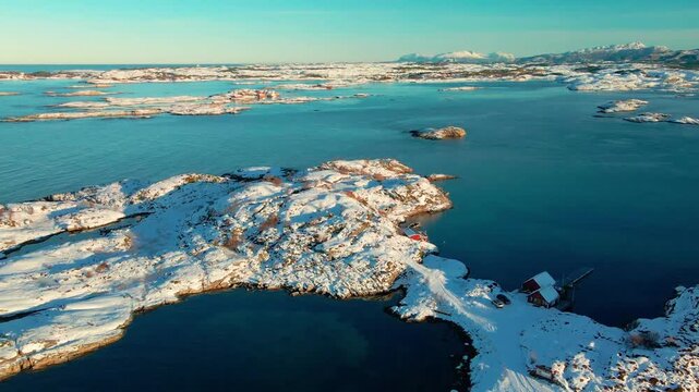 aerial drone shot captures a pristine winter scene in Norway, featuring snow covered islands and rugged coastline meeting the deep blue ocean.