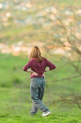 A fair-skinned woman, facing away from the viewer, pauses on a grassy hill at twilight. She rests...