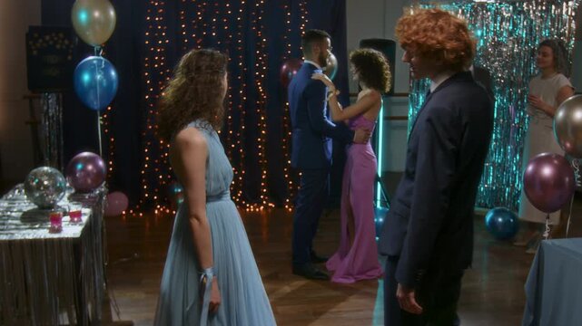 Young red-haired man in formal suit inviting his girfriend, wearing evening dress, to slow dance at high chool prom party