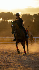 A young woman galloping on horseback at sunset with the golden hour sunlight illuminating her from behind, creating a silhouette on a dusty riding track.