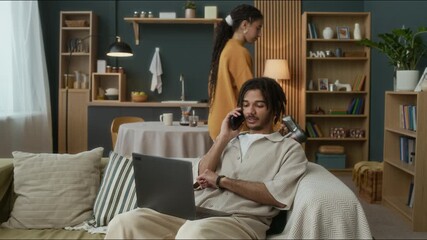 Concentrated young biracial guy sitting on couch working on laptop while talking on phone with his girlfrind or wife vacuuming floor doing household chores - Powered by Adobe