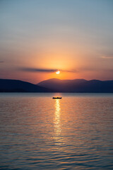 Obraz premium Tranquil fishing boat at sunset over Tolo Bay Greece. Glowing orange sky and distant mountain silhouettes reflecting on calm sea water