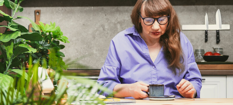 Elderly business woman drinking coffee from blue cup while working at desk in stylish modern apartment - Powered by Adobe