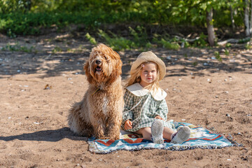 A curly-haired brown Labradoodle or Cavapoo dog with a little child girl in dress are best friends by the sea.