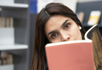 Woman reading a book, peeking over its cover, library setting