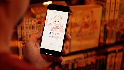 Reading anime maid girls digital manga comic on smartphone beside a shelf with books over shoulder...
