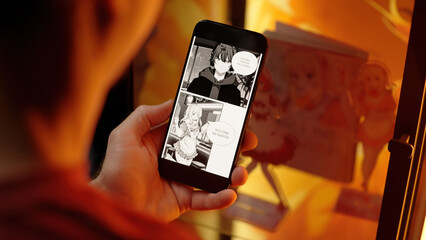 Reading cute anime maid girls manga comic on smartphone beside a merchandise shelf with figurines over shoulder view