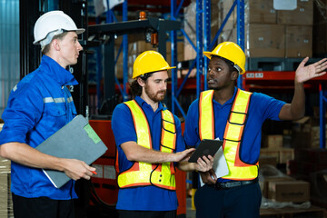 Multiracial group of three warehouse workers standing together, engaged in serious discussion during logistics operation, holding tablet and clipboard, wearing hard hats and safety vests