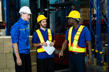 Multiracial group of male adult warehouse workers discussing logistics task near forklift holding paper and tablet wearing uniform and helmet during teamwork coordination inside storage zone