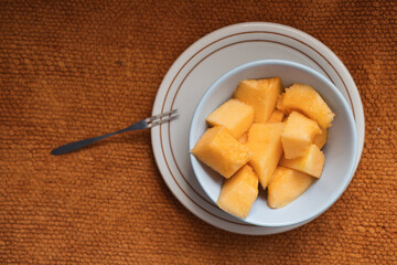 Fresh melon cubes served in a white bowl on a textured orange placemat with a silver fork next to the bowl