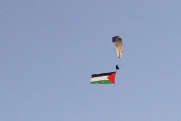 A paraglider flies in a clear blue sky, carrying a large Palestinian flag. An example of a story about freedom, human rights, protest and hope. A sense of freedom and national pride.