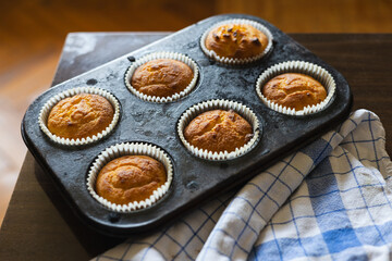 Freshly baked muffins cooling in a black tin on a wooden countertop beside a blue and white checkered cloth