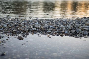 Calm riverbank with smooth pebbles and gentle water reflections during sunset near a serene landscape