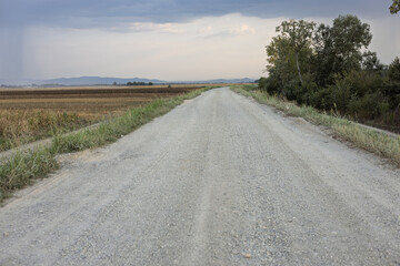 Dusty gravel road stretches across open fields under a cloudy sky at sunset, inviting exploration in a serene landscape