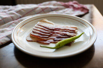 Thinly sliced cured meat arranged elegantly on a plate with a fresh green pepper, showcasing...