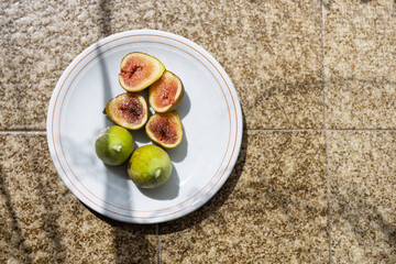 Fresh figs and green lemons on a white plate placed on textured flooring in bright sunlight during midday
