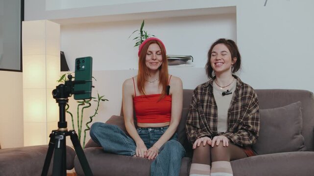 Two girls sit on a sofa with eyes closed, playfully pointing at each other during a fun guessing game, creating lively and engaging content for their audience