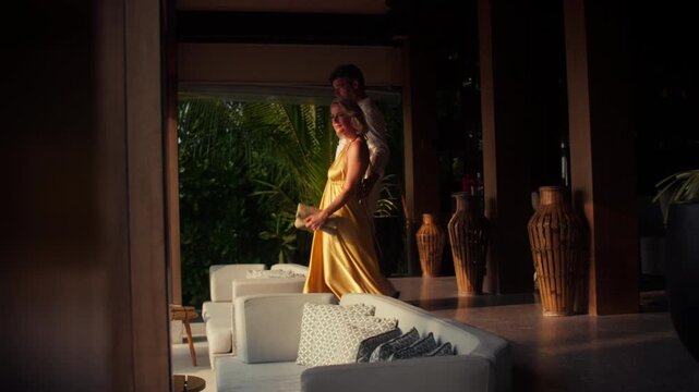 An elegant couple dressed formally walks through a luxurious, open-air resort lounge. They pause to admire the beautiful tropical evening, embodying relaxation and a high-end vacation lifestyle.