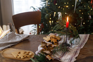 Christmas gingerbread cookies on plate, fir branches, candle and metal cutters on rustic table against decorated christmas tree in evening. Holiday preparation, atmospheric time