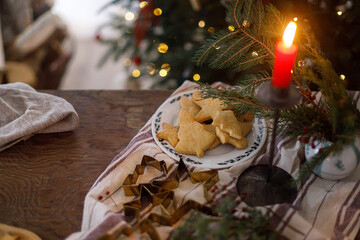 Making holiday cookies. Christmas gingerbread cookies on plate, fir branches, candle and metal cutters on rustic table against decorated christmas tree in evening. Holiday preparation