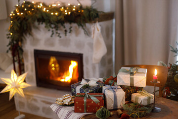 Merry Christmas! Stylish wrapped christmas presents, ornaments, candle, gingerbread cookies on table on background of festive decorated fireplace and christmas lights. Holiday eve