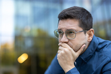 Obraz premium Pensive man wearing glasses contemplating outdoors against a blurred urban backdrop