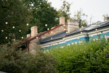 Fototapeta premium Outdoor string lights glowing above traditional house rooftops with brick chimneys and greenery, peaceful garden scene showing cozy decoration and urban evening lifestyle