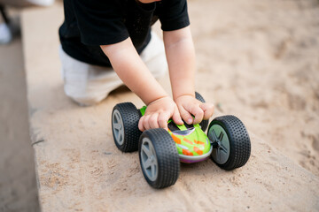 Toddler in sandbox playing with colorful toy car, hands pushing vehicle on sand surface, symbolizing early childhood play, learning, imagination, development and fun