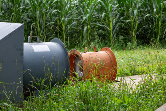 Old Cylindrical Metal Grain Aeration Fan Farm Equipment Among Tall Corn Stalks - For Crop Conditioning in Storage Bin