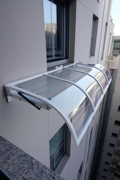 Cubre tendal moderno con estructura met&aacute;lica blanca y paneles transparentes curvados, instalado sobre ventana en fachada urbana, dise&ntilde;ado para proteger de lluvia y sol con est&eacute;tica y arquitectura