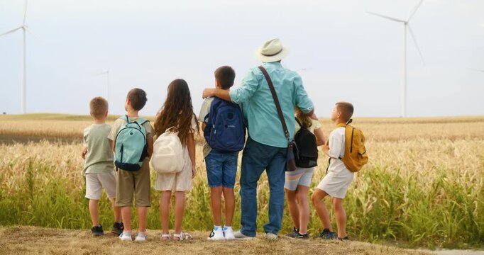 Back view of teacher with young students on meadow near wind turbines, observing and learning about wind energy, sustainable education in nature