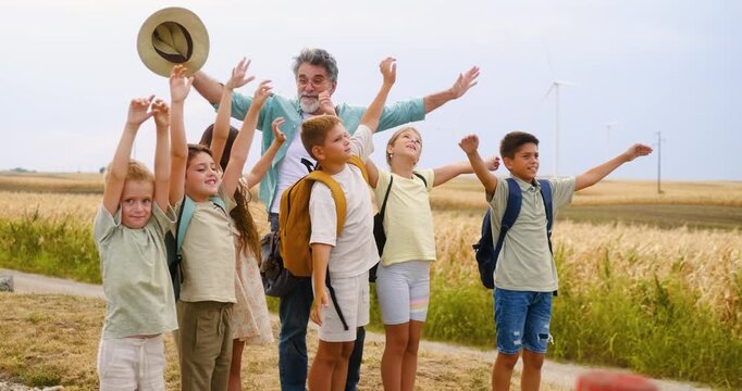 Elderly eacher with group of school children smiling and raising hands while looking into the distance during an outdoor class in nature, enjoying group activity.