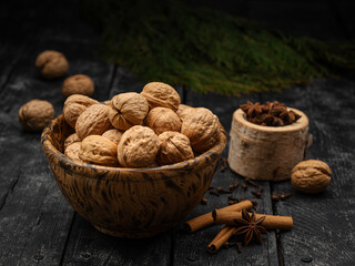 healthy whole walnuts in shell with cinnamon sticks and star anise spice in a wood bowl on a dark and moody black plank table scene. Greenery pine branch.