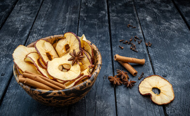 healthy dried dehydrated apple snack slices with cinnamon sticks and star anise spice in a wood bowl on a dark and moody black plank table scene. 