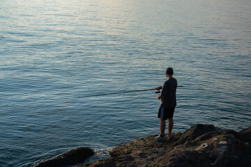 Silhouette of young man fishing at sunrise by sea. Fisherman casting rod against rising sun. Peaceful scene at dawn on coast