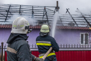 Fototapeta premium Two firefighters spray water on a house's partially collapsed roof, working to extinguish a fire. The image, taken from behind, highlights their teamwork and the ongoing emergency response