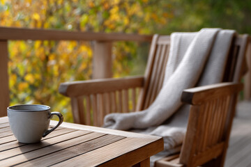 Fototapeta premium Cozy balcony terrace in sunny autumn day. Cup of coffee tea on wooden table, soft blanket draped over a chair